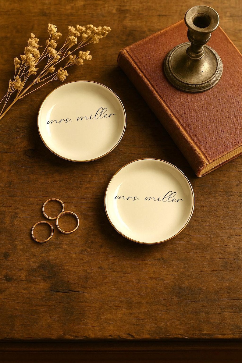 May include: Two small, off-white ceramic dishes with gold rims, each inscribed with "mrs. miller" in black script. Three gold rings and a vintage candlestick holder sit on a wooden surface, alongside a closed book and dried flowers.