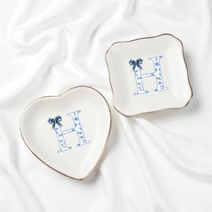 May include: Two decorative trinket dishes. One is heart-shaped, the other is square with scalloped edges. Both are white with a gold trim and feature a blue floral "H" design and a blue bow.