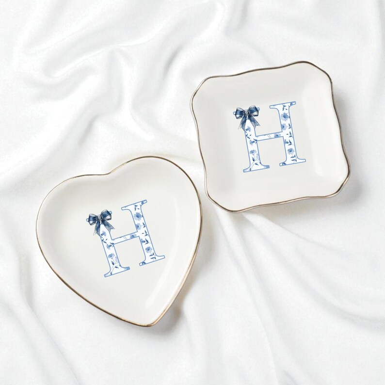 May include: Two decorative trinket dishes. One is heart-shaped, the other is square with scalloped edges. Both are white with a gold trim and feature a blue floral "H" design and a blue bow.
