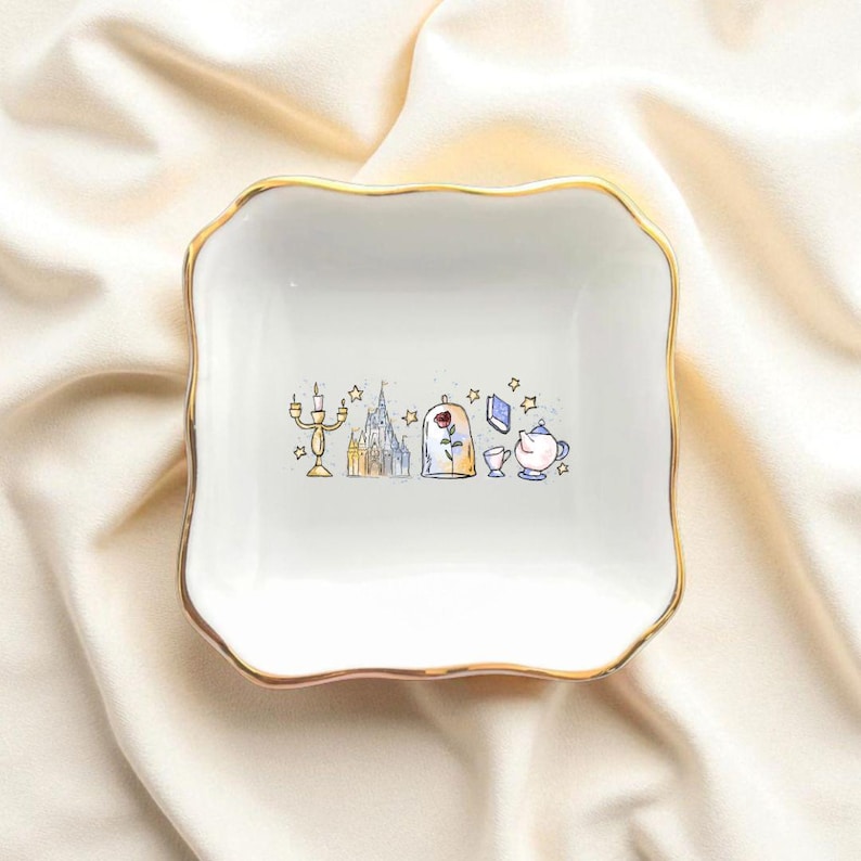 May include: A white, square trinket dish with a gold scalloped edge. The dish features a whimsical illustration of a castle, rose, and teacup. The design is in soft watercolor tones, creating a charming and delicate aesthetic.