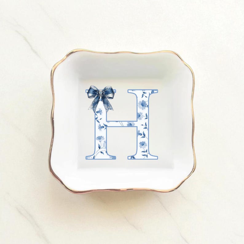 May include: Square white trinket dish with a gold rim. The dish features a blue floral letter "H" and a blue bow. The letter is decorated with blue flowers and leaves. The dish is likely ceramic.