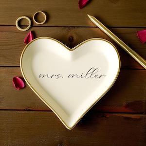 May include: A heart-shaped, off-white ceramic ring dish with a gold rim and the inscription "mrs. miller" in black script. Two gold rings and a gold pen are also visible, along with scattered red rose petals, all on a dark wood surface.