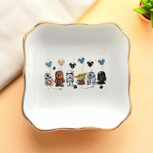 May include: White ceramic trinket dish with a gold rim, featuring Star Wars characters holding balloons. The characters include BB-8, Chewbacca, a Stormtrooper, Grogu, R2-D2, and Darth Vader. The dish has a scalloped edge.