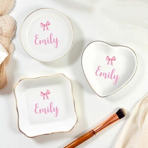 K&ouml;nnte beinhalten: Drei wei&szlig;e Keramik-Schmuckschalen mit goldenen R&auml;ndern. Jede Schale hat eine rosa Schleife und den Namen "Emily" in Rosa aufgedruckt. Eine Schale ist rund, eine herzf&ouml;rmig und eine quadratisch mit gewellten Kanten. Ein Make-up-Pinsel ist im Vordergrund.