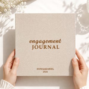 Puede incluir: Un diario de compromiso cuadrado con cubierta de lino beige. La parte frontal presenta el texto en marrón engagement JOURNAL, seguido por los nombres SOPHIA&DANIEL y el año 2026 impresos en una fuente más pequeña y clara centrada abajo.