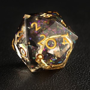 May include: A clear, multi-faceted D20 die with gold-colored numbering and accents. The die is filled with iridescent glitter and sits on a black surface. The numbers are clearly visible, making it suitable for tabletop gaming.