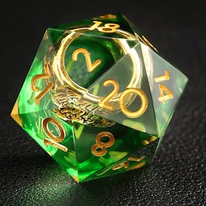 May include: A transparent green twenty-sided die with gold-colored numbers and a gold-colored interior design. The die is designed for tabletop role-playing games. The numbers are clearly visible on each face.