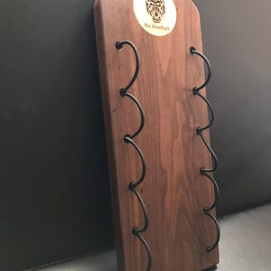 May include: A wooden wine rack with a dark finish, featuring curved metal holders for bottles. The rack has a rounded top with a circular logo that reads "The Silverback". The design is simple and elegant.