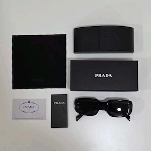 Peut inclure: Lunettes de soleil Prada noires à monture rectangulaire, présentées avec un étui noir, une boîte, un chiffon de nettoyage et une carte d'authentification. Le logo Prada est visible.