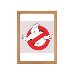 May include: A framed cross-stitch design featuring the Ghostbusters logo. The logo is a red circle with a diagonal line through it, and a white ghost with a surprised expression is inside the circle.