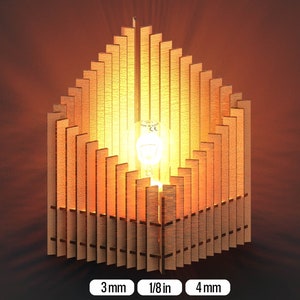 Puede incluir: Una pantalla de lámpara de madera con un diseño geométrico. La pantalla está hecha de finas lamas de madera dispuestas en un patrón triangular. La pantalla está iluminada desde el interior, creando un brillo cálido. La pantalla tiene 10 cm de alto y 7,5 cm de ancho.