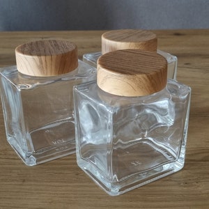 Può includere: Tre barattoli quadrati in vetro trasparente con coperchi in legno. I barattoli sono vuoti e hanno una superficie leggermente ruvida. I coperchi sono di colore marrone chiaro e hanno una venatura del legno.