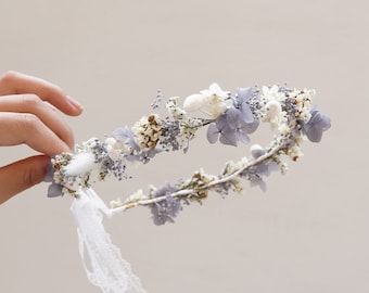 May include: A delicate floral crown made with dried flowers in shades of white and light blue. The crown is adorned with small white flowers, sprigs of dried baby's breath, and larger blue hydrangea blooms. A white lace ribbon is tied at the back of the crown.