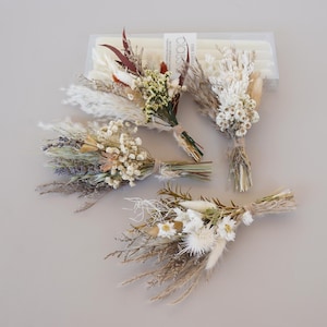 May include: Four small dried flower bouquets in shades of white, beige, green, and purple. Each bouquet is tied with twine. A package of white candles is visible in the background.