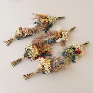 May include: Five small bouquets of dried flowers in various colours, including white, yellow, orange, red, and blue. The bouquets are tied with twine and arranged on a white surface.