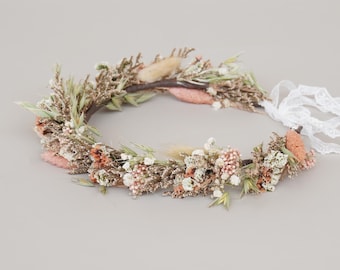 May include: A delicate floral crown made with dried flowers and grasses. The crown features a variety of dried flowers in shades of white, pink, and brown. The crown is tied with a white lace ribbon.