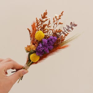 May include: A small bouquet of dried flowers in shades of orange, yellow, and purple. The bouquet includes dried grasses, small yellow pom poms, and purple statice flowers.