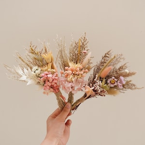 May include: Four small bouquets of dried flowers in shades of pink, white, and beige. The bouquets are tied together with twine and feature dried grasses and pampas grass.