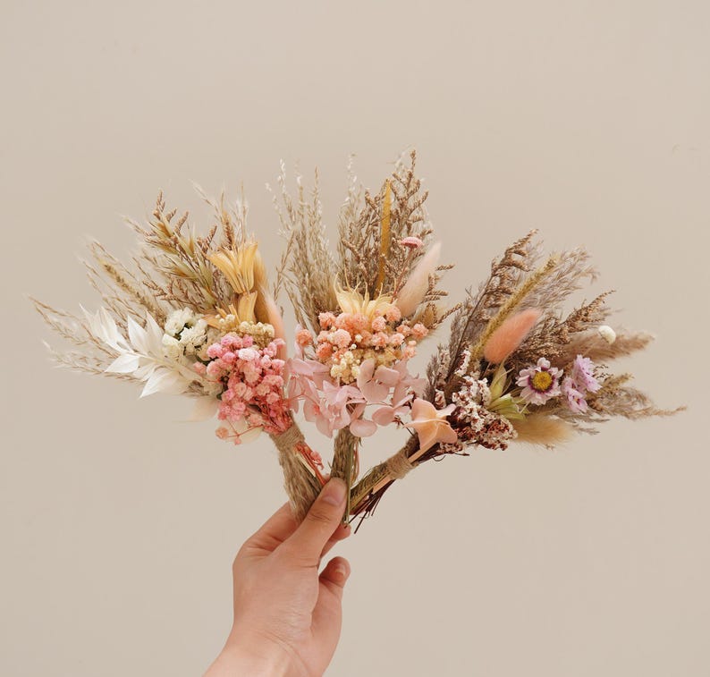 May include: Four small bouquets of dried flowers in shades of pink, white, and beige. The bouquets are tied together with twine and feature dried grasses and pampas grass.