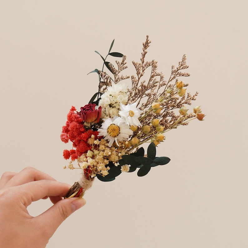 May include: A small bouquet of dried flowers, including white daisies, red and orange flowers, and brown dried grasses. The bouquet is tied together with twine.