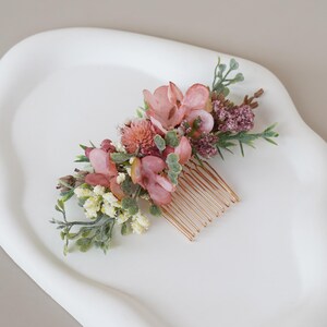 Könnte beinhalten: Ein floraler Haarkamm mit rosa und malvenfarbenen Blüten, grünen Blättern und kleinen weißen Blüten. Die Blumen sind auf einem goldfarbenen Kamm angeordnet und schaffen ein zartes und elegantes Accessoire. Dieser Haarkamm ist ideal für Hochzeiten oder besondere Anlässe.