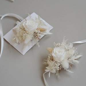May include: Two white dried flower boutonnieres with white roses and dried grasses. The boutonnieres are tied with white ribbon.