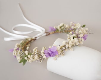 May include: A delicate floral crown with white baby's breath and purple flowers. The crown is made with a wire base and has a white ribbon tie.
