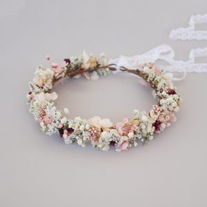 Può includere: Una delicata corona floreale realizzata con fiori secchi in tonalità di bianco, rosa e bordeaux. La corona è realizzata con una base naturale e rustica ed è legata con un nastro bianco.