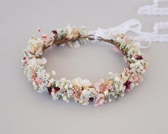 May include: A delicate floral crown made with dried flowers in shades of white, pink, and burgundy. The crown is made with a natural, rustic base and is tied with a white ribbon.