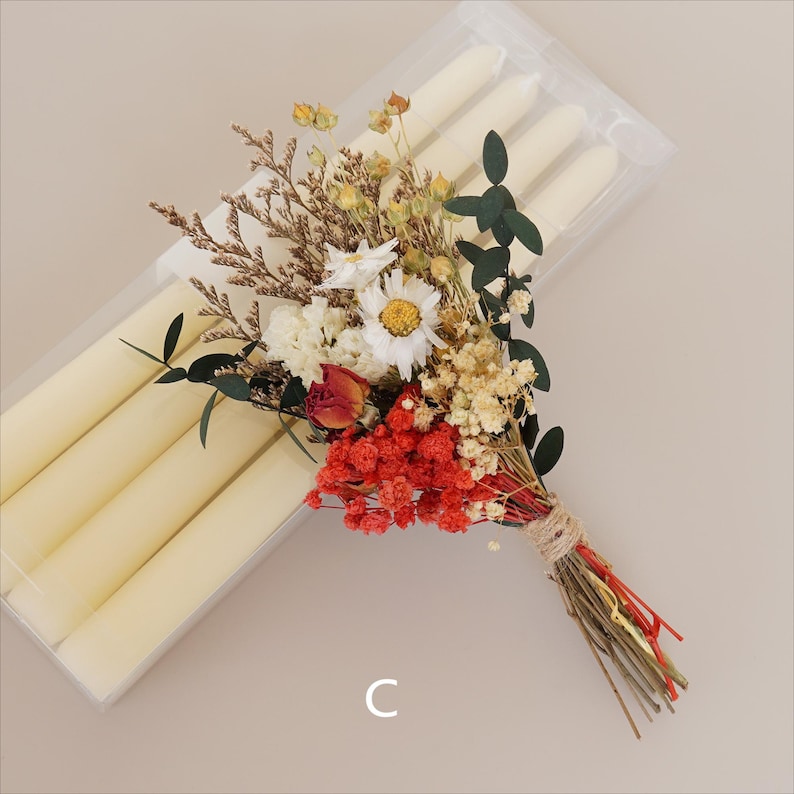 May include: A bouquet of dried flowers in shades of white, red, and yellow, tied with twine. The bouquet is next to a clear plastic package of white taper candles.
