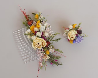 May include: A floral hair comb and matching boutonniere featuring dried flowers in shades of yellow, white, red, and purple. The comb has a silver, plastic base. The boutonniere is a small, round arrangement of similar flowers.