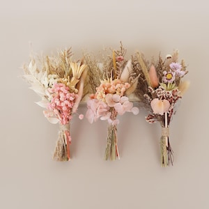 May include: Three dried flower bouquets with pink, beige, and brown tones. The bouquets are tied with twine and are hanging from a white wall.