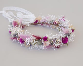 May include: A delicate floral crown made with dried flowers. The crown is made with white baby's breath, pink and purple wildflowers, and dried grasses. The crown is tied with a white lace ribbon.