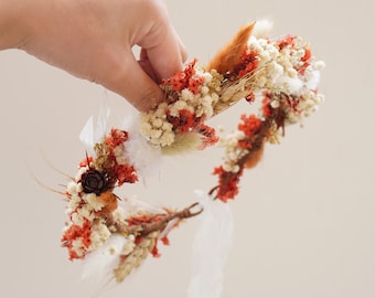 May include: A dried flower crown with white, orange, and red flowers, and brown stems. The crown is made with natural materials and has a rustic, bohemian style.