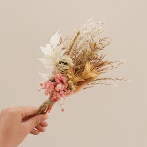 May include: A small bouquet of dried flowers in shades of pink, white, and brown. The bouquet includes dried grasses, pampas grass, and small pink flowers.