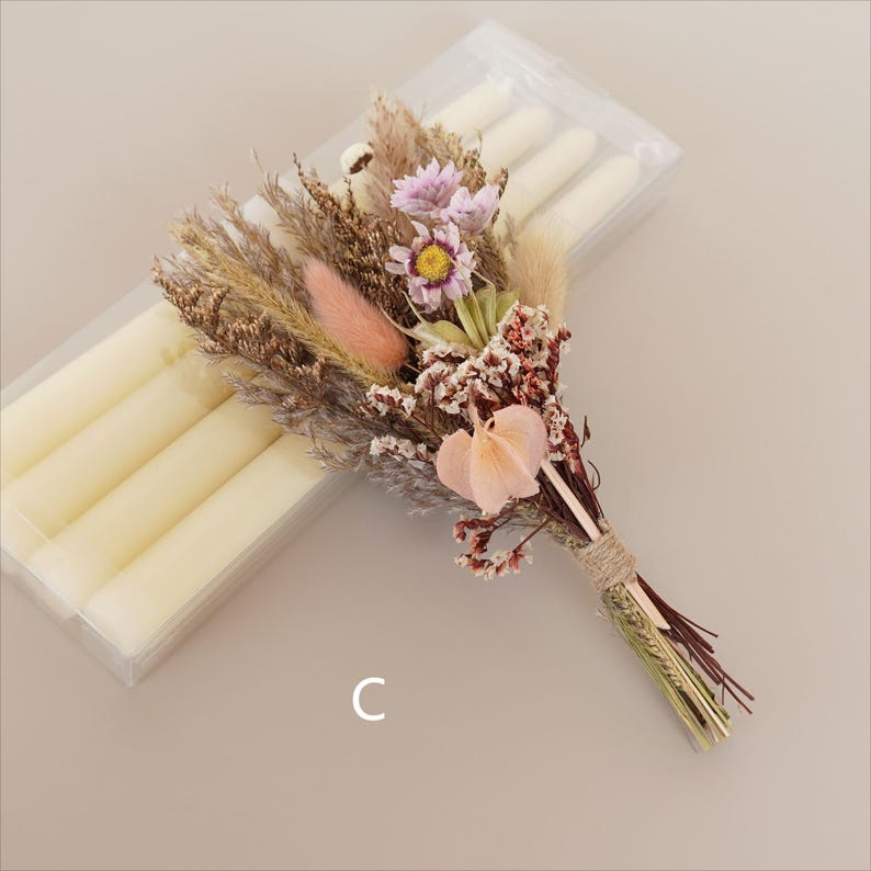 May include: A bouquet of dried flowers in shades of pink, brown, and white, tied with twine. The bouquet is next to a package of white taper candles.
