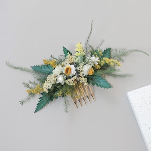 Könnte beinhalten: Ein goldfarbener Haarkamm, verziert mit einer Ansammlung getrockneter Blumen in Weiß, Gelb und Grün. Das Blumenarrangement umfasst Gänseblümchen und andere Wildblumen und schafft einen natürlichen, eleganten Look. Die Zähne des Kamms sind so konzipiert, dass sie die Blumen im Haar befestigen.