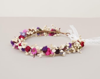 May include: A floral crown made with dried flowers in shades of pink, purple, and red. The crown is made with a wire base and has a white ribbon attached.