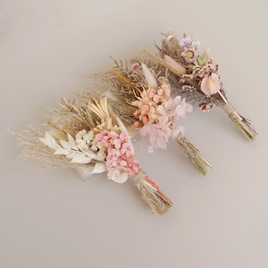 May include: Three small bouquets of dried flowers in shades of pink, beige, and white. The bouquets are tied with twine and feature dried grasses, pampas grass, and other dried flowers.