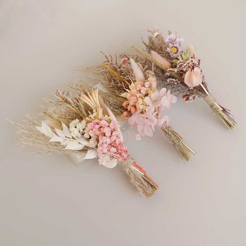 May include: Three small bouquets of dried flowers in shades of pink, beige, and white. The bouquets are tied with twine and feature dried grasses, pampas grass, and other dried flowers.