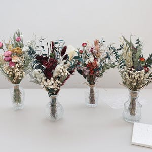 May include: Four bouquets of dried flowers in clear glass vases. The bouquets feature a variety of colors, including pink, red, white, and green. Each bouquet is tied with twine. The vases are arranged on a light gray surface.