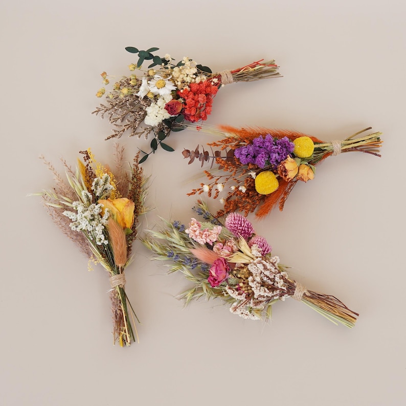 May include: Four small bouquets of dried flowers in various colors, including yellow, orange, purple, pink, and white. The bouquets are tied with twine and are arranged on a light brown background.