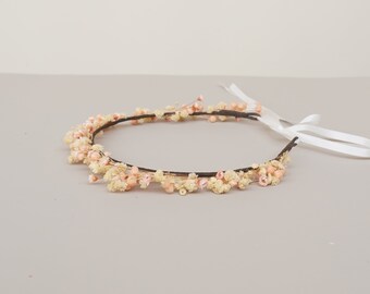 May include: A delicate floral crown made with dried flowers in shades of white and pink. The crown is made with a wire base and has a white ribbon for tying.