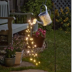 May include: A solar-powered garden light shaped like a watering can, featuring a floral design. It hangs from a black metal stake, with a string of warm white LED lights resembling water. The scene is set in a garden.