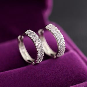 Peut inclure: Une paire de boucles d'oreilles créoles en argent, serties de petites pierres claires et scintillantes. Les boucles d'oreilles sont présentées sur un fond de velours violet foncé, créant un contraste qui met en valeur leur éclat. Bijou classique.
