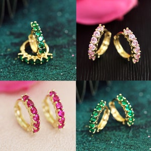 May include: Four pairs of gold hoop earrings with different coloured gemstones. The earrings are on a variety of surfaces, including green, pink, and white.