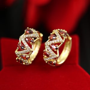 May include: A pair of gold hoop earrings with a geometric design featuring red and white stones.