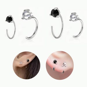 Può includere: Orecchini a cerchio argentati con un design curvo. Ogni orecchino presenta una piccola gemma rotonda, con coppie che mostrano una pietra trasparente o nera. L'immagine include anche primi piani degli orecchini indossati.