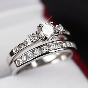 Puede incluir: Un par de alianzas de plata con un gran diamante central brillante y diamantes más pequeños en la banda. Los anillos están en una caja blanca.