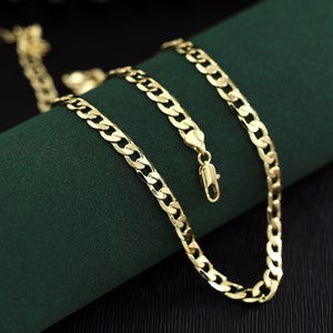 May include: A gold chain necklace with a curb link design. The chain is lying on a green surface.